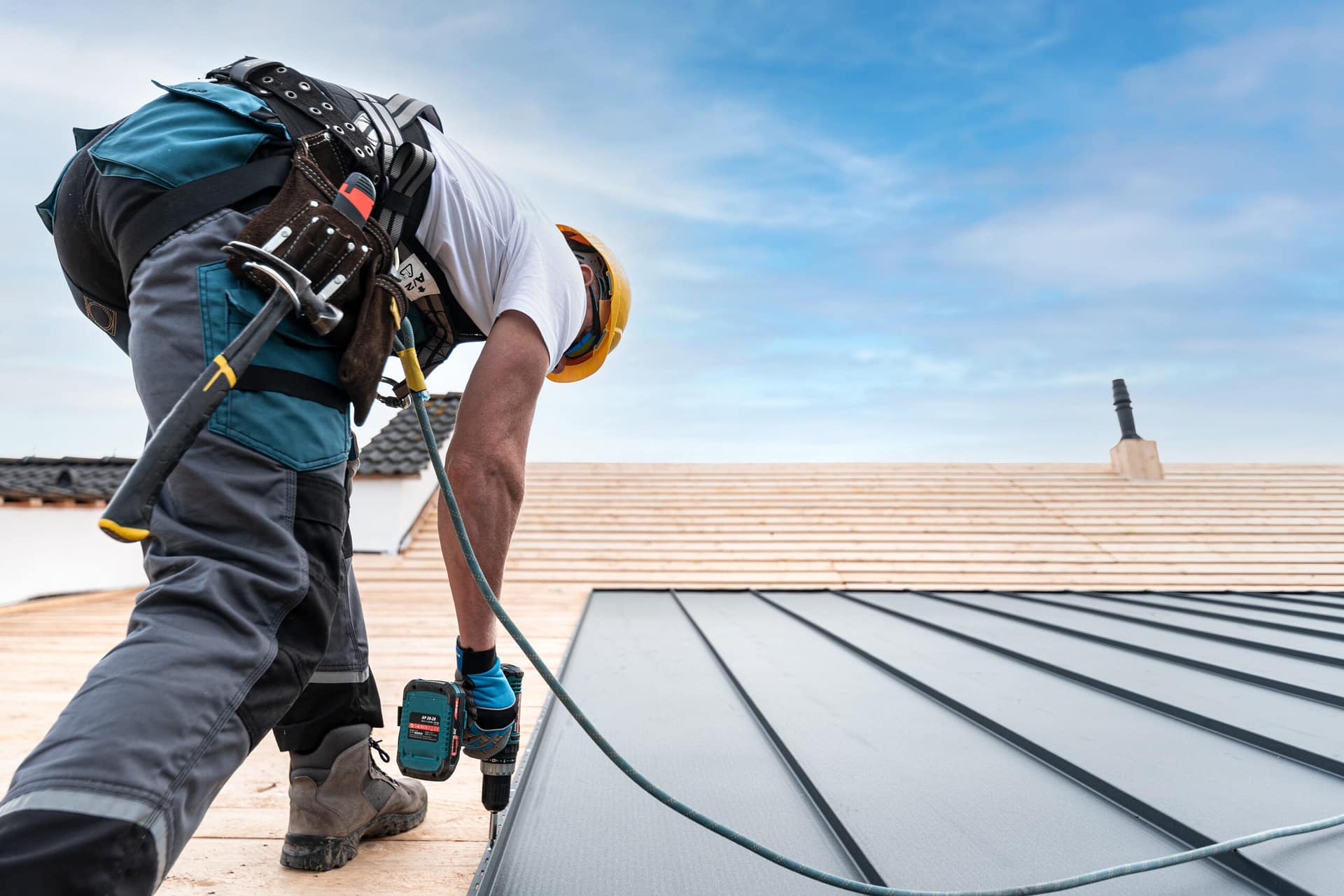 A roofer with a safety harness and tool belt is working with a e Roofer installing Metal Roof